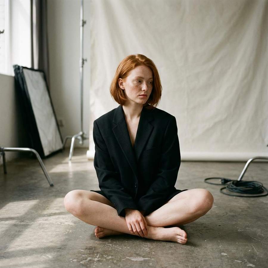 AI-generated fashion photograph of a red-haired woman in a black blazer seated in a photo studio